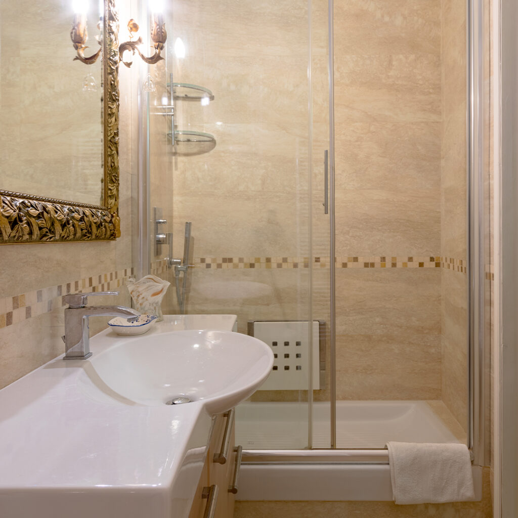 Il bagno è dotato di un’ampia doccia che garantisce comfort e praticità, oltre a un grande specchio impreziosito da una cornice intarsiata in legno dorato. Il lavandino, dal design moderno e funzionale, completa l’ambiente con un tocco di eleganza.