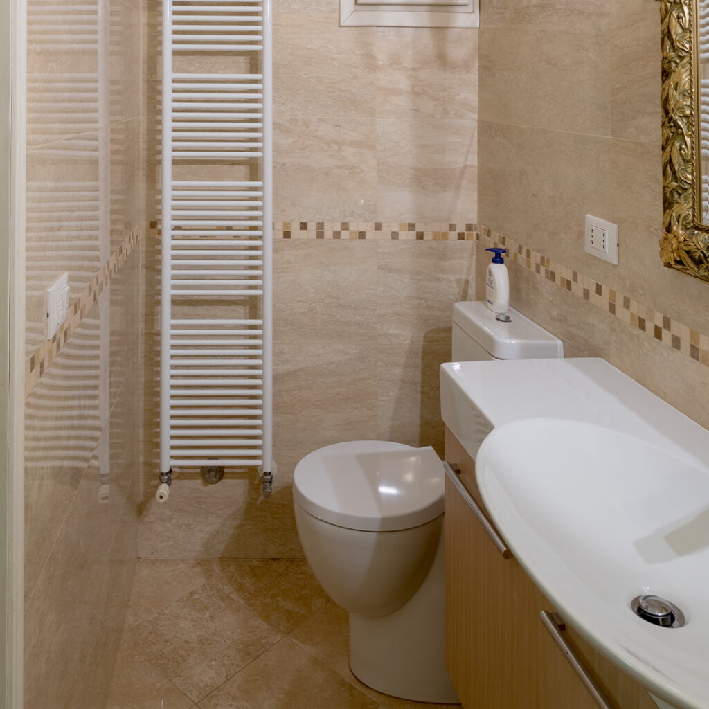 Il bagno, interamente rivestito di eleganti piastrelle beige, offre un’atmosfera calda e accogliente. Un pratico scaldasalviette, che funge anche da sistema di riscaldamento, aggiunge comfort e funzionalità all’ambiente.