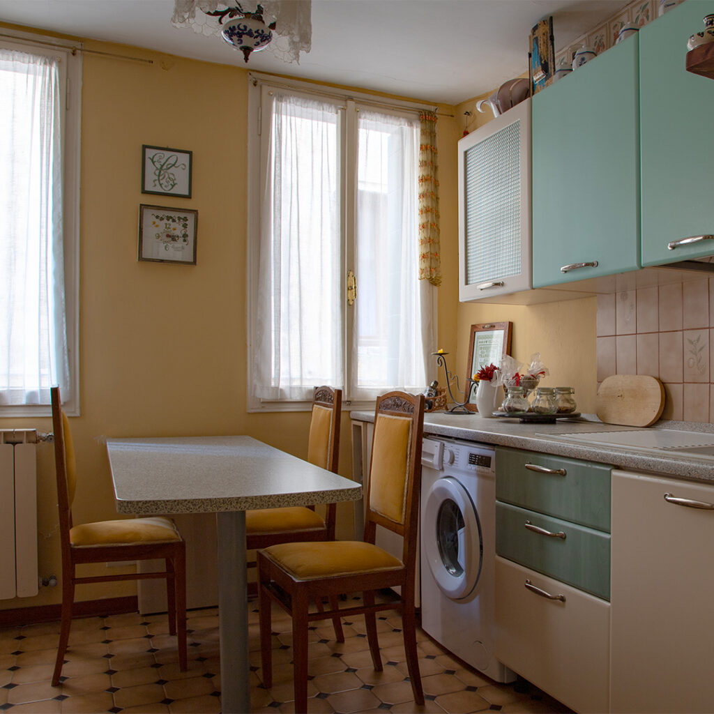 Un accogliente tavolo a isola con sedie in legno imbottite domina l'ambiente, mentre una cucina lineare dal fascino vintage, caratterizzata da ante nei toni del verde e del beige, completa l’arredamento con stile e funzionalità.