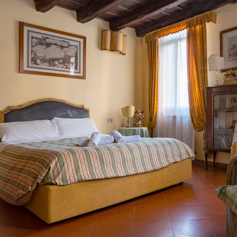 La camera da letto dell'appartamento veneziano è un raffinato connubio di eleganza e storia, arredata con mobili d'antiquariato. Le tende e le pareti, in caldo colore ocra, si armonizzano perfettamente con il pavimento in cotto e le travi in legno a vista che impreziosiscono il soffitto, creando un’atmosfera autentica e accogliente.